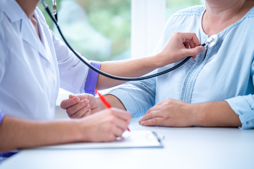 Eine Ärztin oder ein Arzt hört die Brust einer Patientin mithilfe eines Stethoskops ab. Dabei macht sie Notizen auf einem Block.