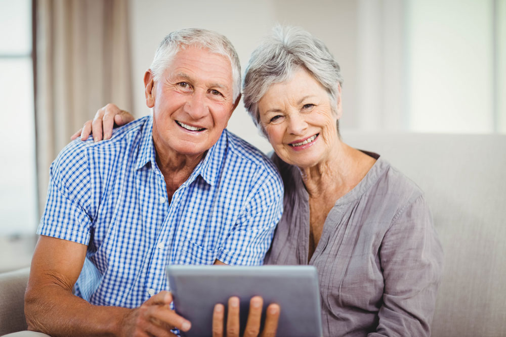 Ein älteres Pärchen sitzt auf einem grauen Sofa und lächelt in die Kamera, in der Hand halten sie ein Tablett.