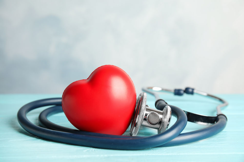 Ein Stethoskop liegt aufgerollt auf einem türkisen Untergrund, das Bruststück berührt ein klassisches, rotes Herz.